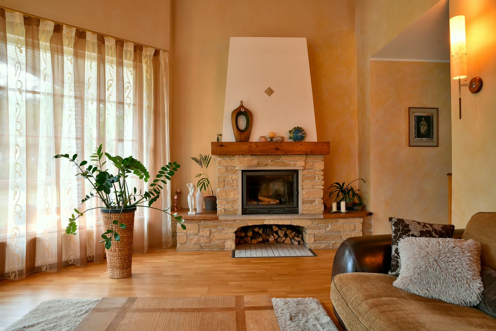 Living room with a stone fireplace, wood floor, a large potted plant, sheer curtains, and a leather sofa in warm afternoon light