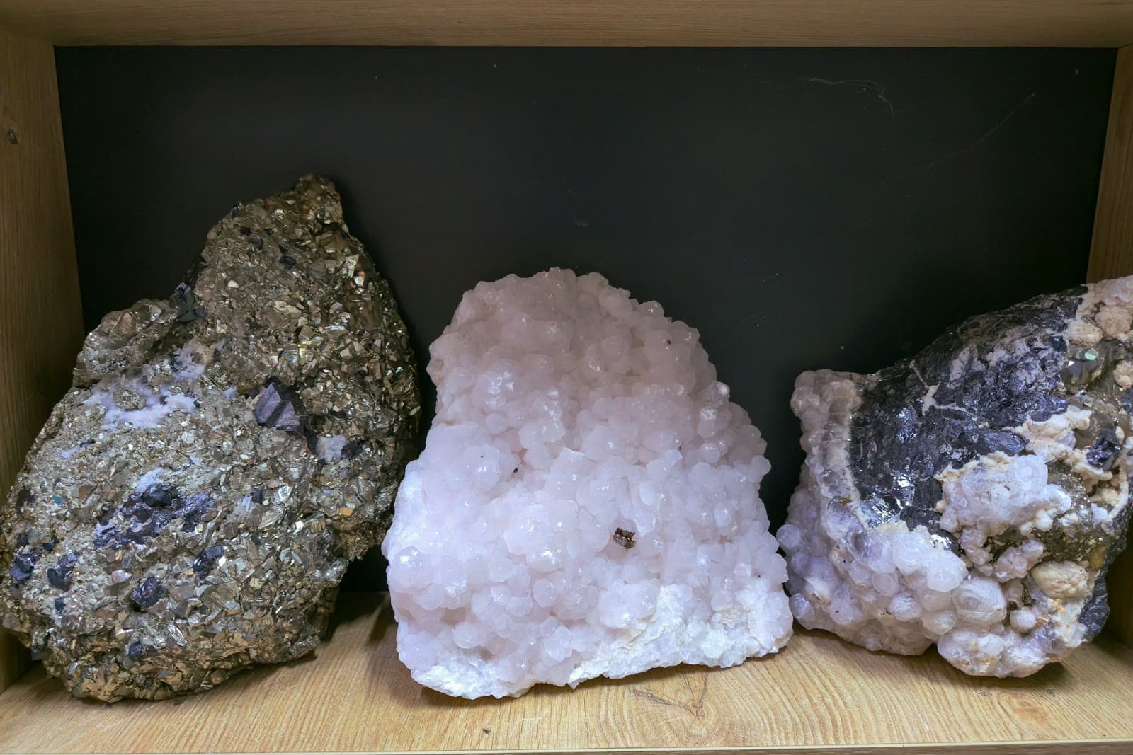 Three raw mineral clusters, including pyrite and white calcite, sitting on a wooden shelf