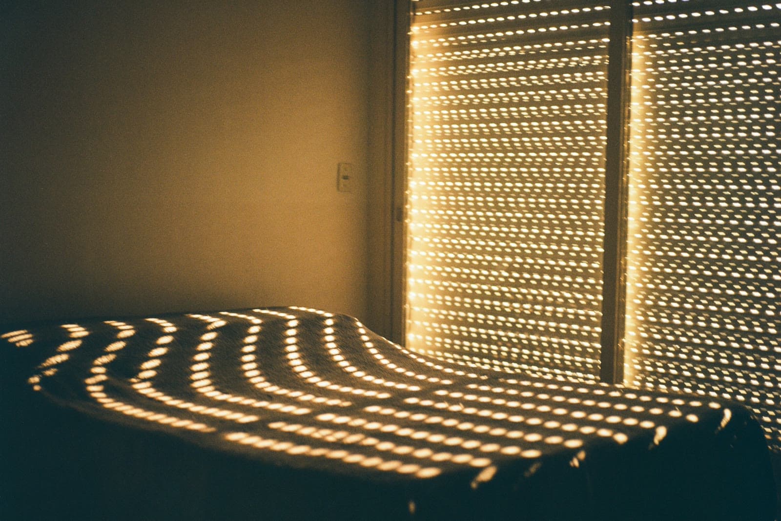 Warm sunlight through a perforated window shutter casting rows of light and shadow across a bed and wall