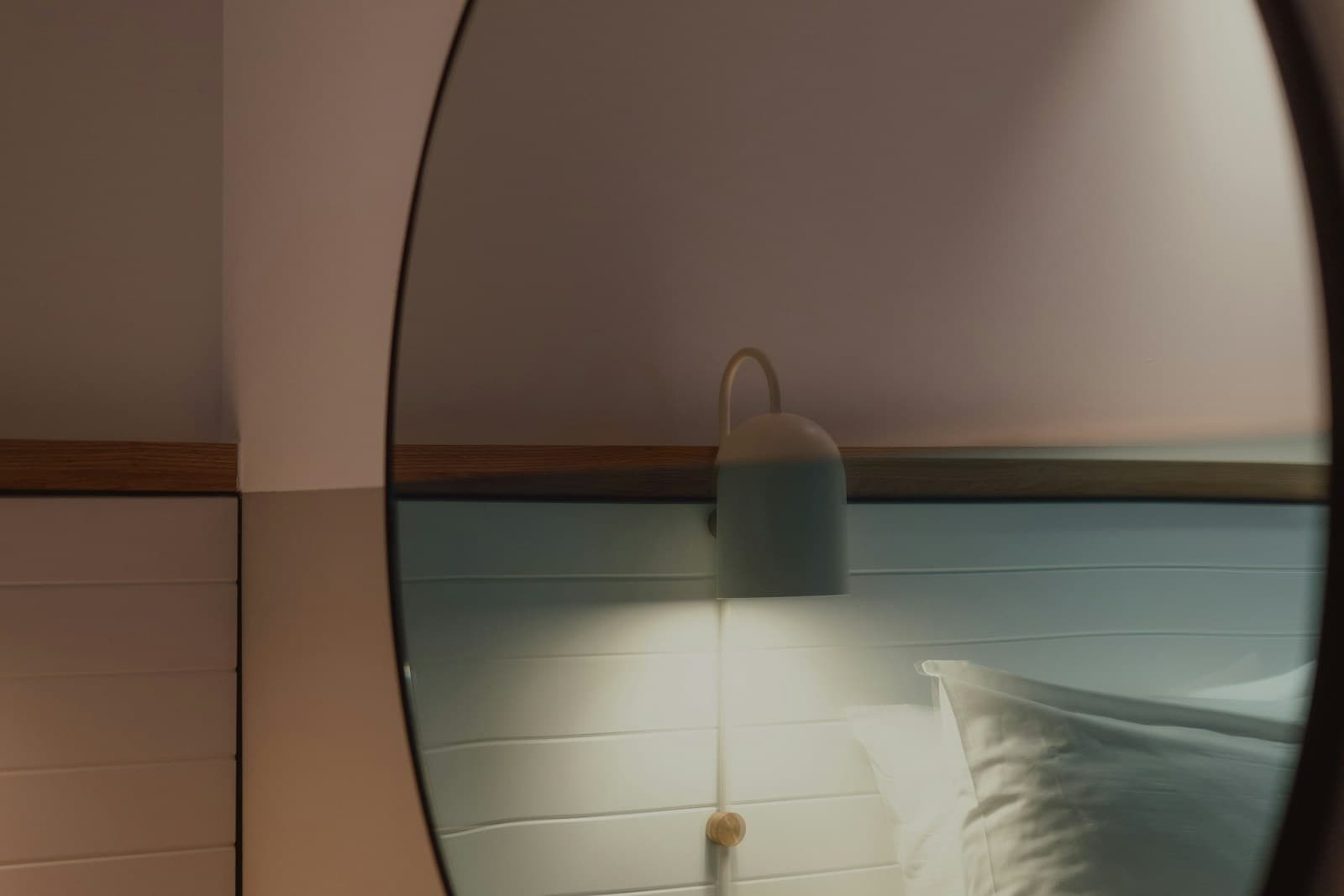 Round mirror reflecting a bedside lamp and pillow in soft evening light