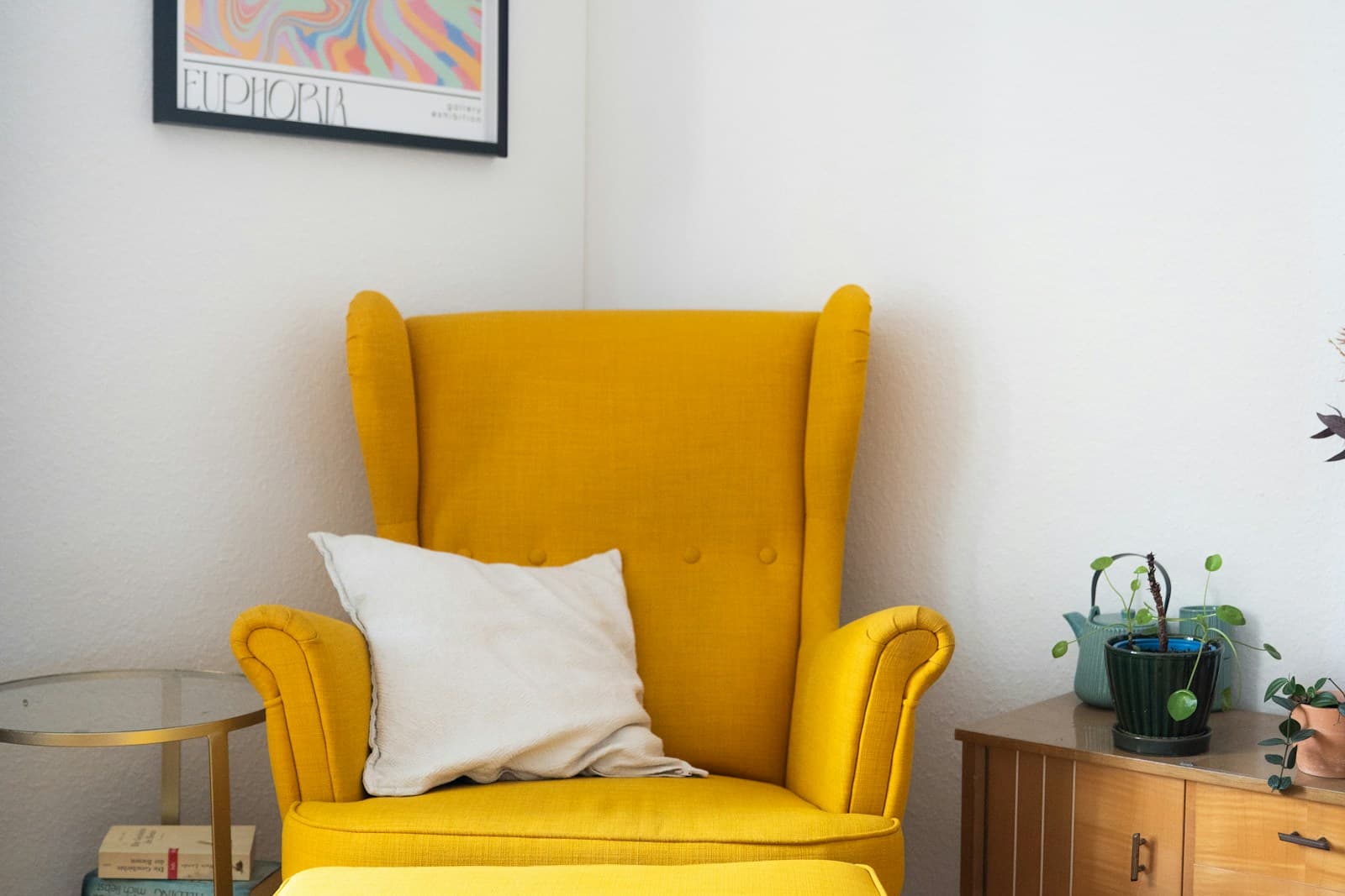 Cozy mustard-yellow wingback chair with a linen pillow, books, and plants in a bright corner